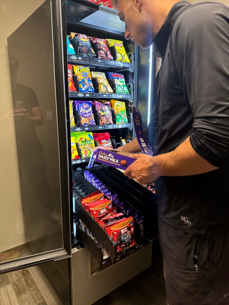 Man in black shirt selecting snack from refrigerated vending machine stocked with colorful packaged snacks